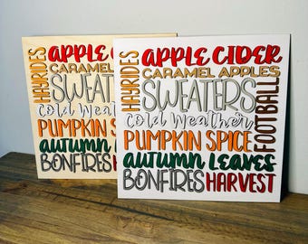 May include: Two square wooden signs featuring layered, colorful text including Apple Cider, Caramel Apples, Sweaters, Cold Weather, Pumpkin Spice, Autumn Leaves, Bonfires, Harvest, Hayrides, and Football. The signs have a white or light wood background.