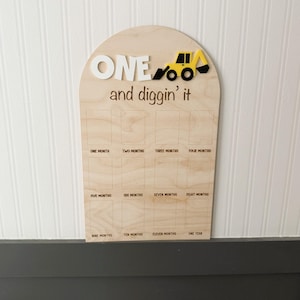 May include: An arched wooden milestone board featuring the white text ONE, a yellow and black construction vehicle icon, and the brown text and diggin' it. Below are twelve rectangular photo slots labeled from one month to one year.