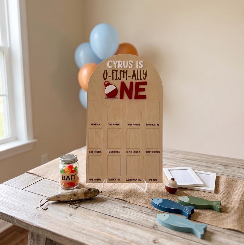May include: An arched wooden milestone board with the text Cyrus is O-Fish-Ally One, featuring twelve rectangular photo slots labeled from newborn to one year. Includes a glass jar labeled Bait, a fishing lure, and three wooden fish cutouts.