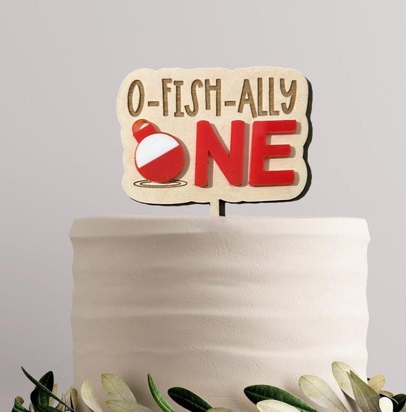 May include: A wooden cake topper with a light tan finish featuring the text O-FISH-ALLY ONE in brown lettering. The letter O is replaced by a red and white fishing bobber graphic. The topper has a single stake for insertion into a cake.