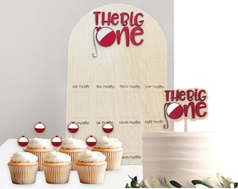 May include: A light wood arched milestone board with the text "THE BIG ONE" and a fishing bobber graphic, featuring twelve rectangular slots labeled by month. Includes a matching cake topper and six small cupcake toppers with bobbers.