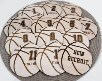 May include: A set of twelve wooden discs, each designed to resemble a basketball. The discs are light-colored with brown etched lines and numbers from 1 to 11, plus one that says "NEW RECRUIT". The discs are arranged on a gray circular mat.