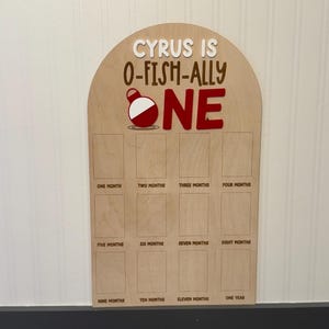May include: An arched wooden milestone board featuring the text CYRUS IS O-FISH-ALLY ONE with a red and white fishing bobber graphic. Below the text are twelve rectangular photo slots labeled from one month to one year in brown lettering.
