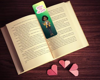 Op de afbeelding: Een bladwijzer met de tekst "NOW OR NEVER" en een illustratie van een vrouw in een blazer, ingevoegd in een open boek. Drie origami-harten liggen naast het boek op een houten oppervlak. De bladwijzer is groen en geel.