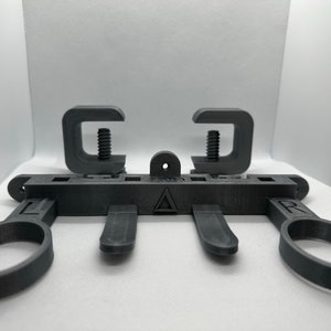 May include: A black 3D printed phone holder with two clamps and a triangular design. The holder has two rings on the sides and three slots in the middle.
