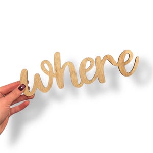 May include: Wooden sign in a cursive font that spells out the word "where". The sign is made of light-colored wood and is held against a white background.