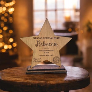 May include: A wooden star-shaped award with the text "SANTAS OFFICIAL STAR" and the name "Rebecca". It also includes the phrases "FOR GOOD BEHAVIOUR IN 2025" and "ON THE NICE LIST!". The award is on a wooden base.