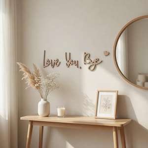 May include: Wooden wall art spelling "Love You, Bye" with a small heart. A round mirror and framed botanical print are on the wall. A wooden table displays a vase with dried flowers, a candle, and a framed picture.