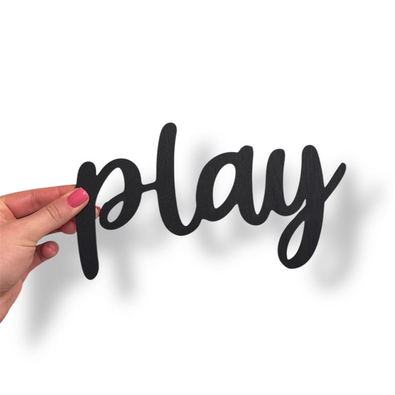 May include: A black wooden sign in a cursive font that spells out the word "play". The sign is held by a hand with pink nail polish against a white background. The sign casts a shadow.