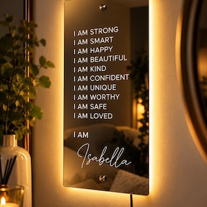 May include: A rectangular wall mirror with integrated warm LED backlighting. The surface features white text: I AM STRONG, I AM SMART, I AM HAPPY, I AM BEAUTIFUL, I AM KIND, I AM CONFIDENT, I AM UNIQUE, I AM WORTHY, I AM SAFE, I AM LOVED, and I AM Isabella.