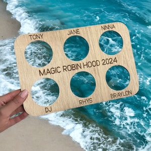 Peut inclure: Planche en bois avec huit découpes circulaires, chacune étiquetée d'un nom. La planche est étiquetée "MAGIC ROBIN HOOD 2024".