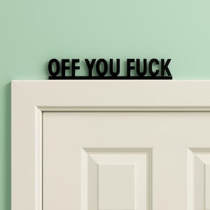 May include: Black wooden sign with the words "OFF YOU FUCK" above a white door. The sign is in a bold, sans-serif font and is against a light green wall.