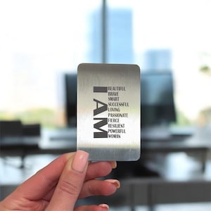 May include: A rectangular, brushed metal card with the word "I AM" in large, bold, black letters on the left side. The right side lists empowering words: Beautiful, Brave, Smart, Successful, Loving, Passionate, Fierce, Resilient, Powerful Woman.