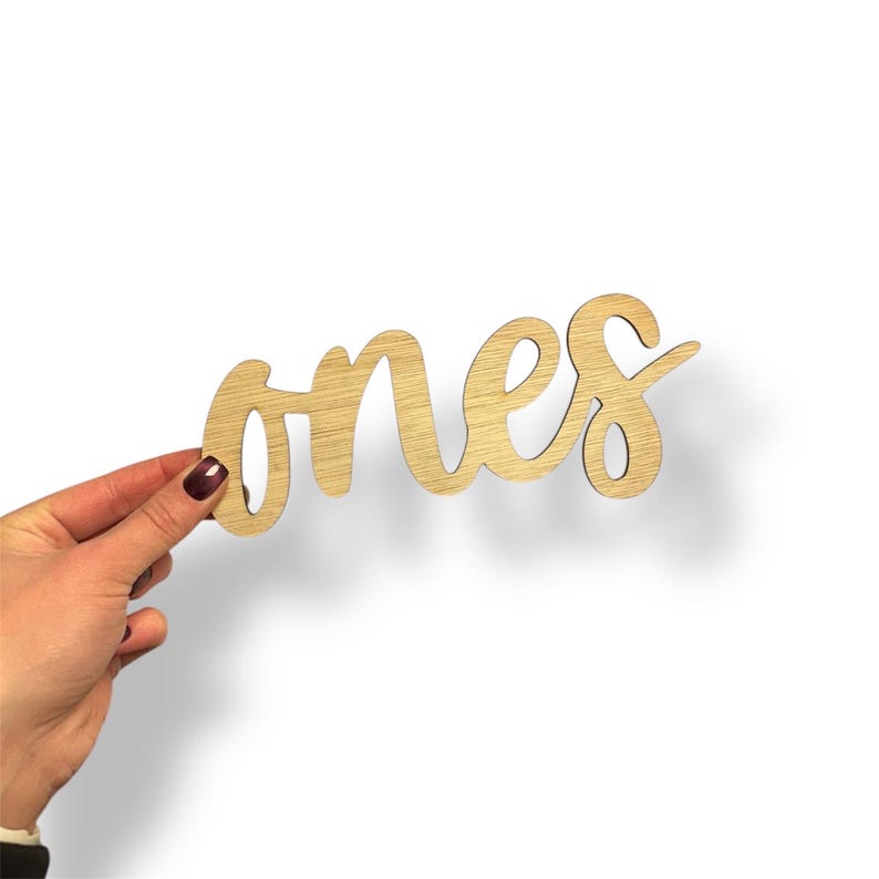May include: Wooden cutout of the word "ones" in a cursive font. The word is held by a hand with a dark red nail polish. The wood has a light brown color and a visible wood grain.