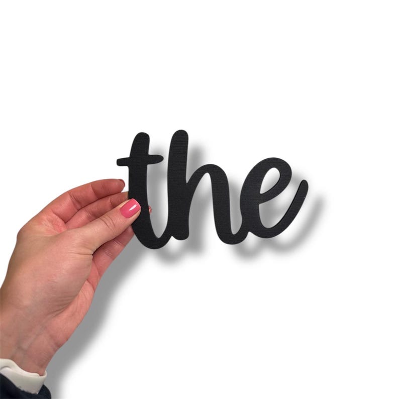 May include: Black wooden cutout of the word "the" in a cursive font. The word is held by a hand with a pink painted fingernail against a white background. The cutout casts a shadow.