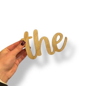 May include: Wooden cutout of the word "the" in a cursive font. The letters are a light brown color, with a visible wood grain. The cutout is held against a white background.