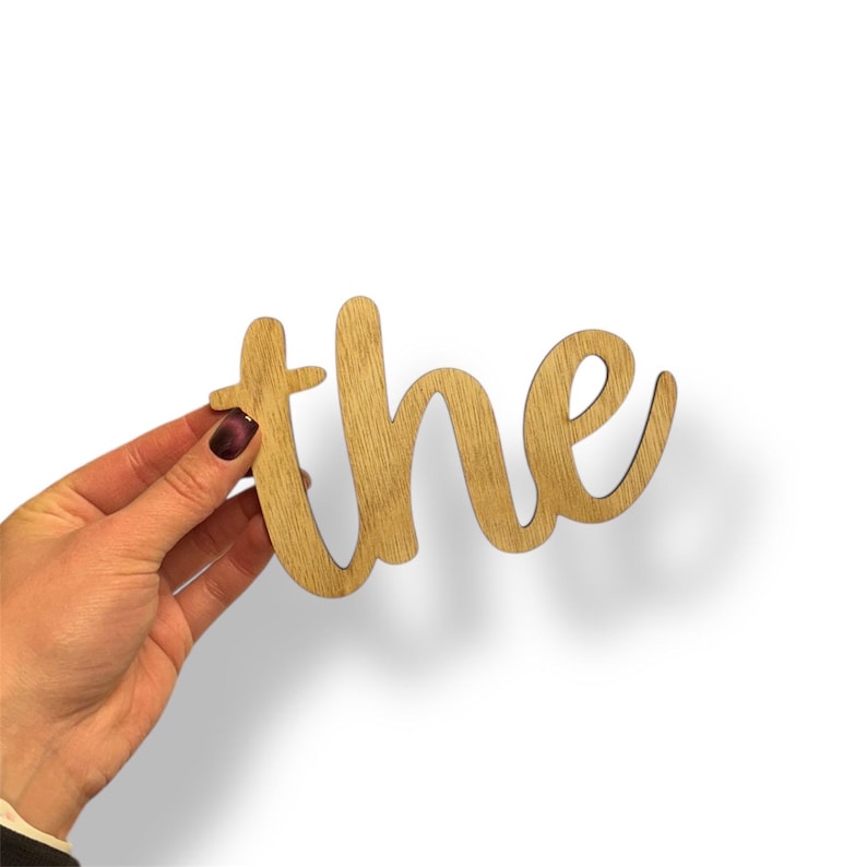 May include: Wooden cutout of the word "the" in a cursive font. The letters are a light brown color, with a visible wood grain. The cutout is held against a white background.