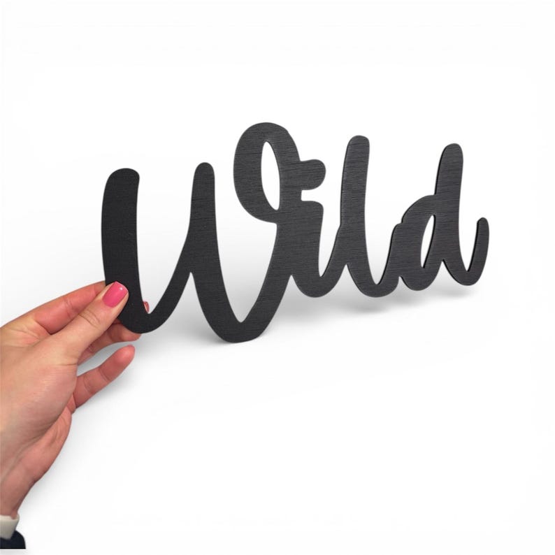 May include: Black wooden sign with the word "Wild" in a cursive font. The sign is held against a white background. The word is cut out of the wood, creating a negative space effect.