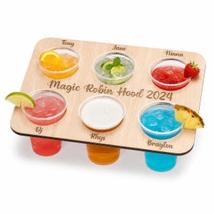 May include: A wooden serving tray with six small cocktails in clear plastic cups. Each drink is a different color and garnished with fruit. The tray is engraved with "Magic Robin Hood 2024" and names for each drink.