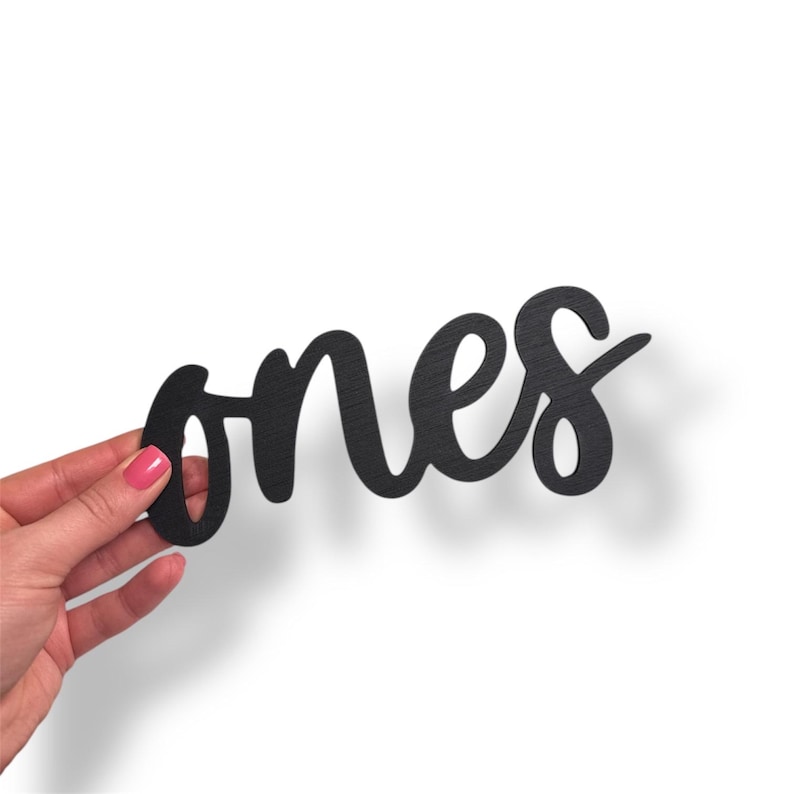 May include: Black wooden sign with the word "ones" in a cursive font. The sign is held by a hand with pink nail polish against a white background. The sign casts a shadow.