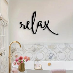 May include: Black cursive "relax" wall art. The word is mounted on a white wall above a bathtub. A gold tap, a vase of pink flowers, and a pink towel are also visible.