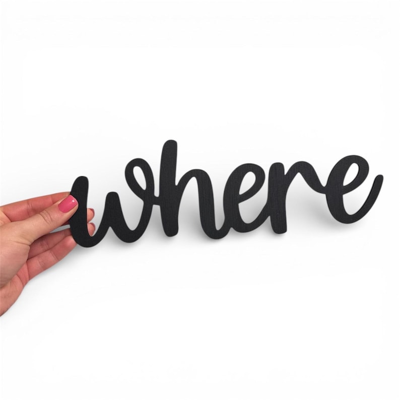 May include: A black wooden sign in a cursive font that spells out the word "where". The sign is held by a hand with pink nail polish against a white background. The sign is a decorative item.