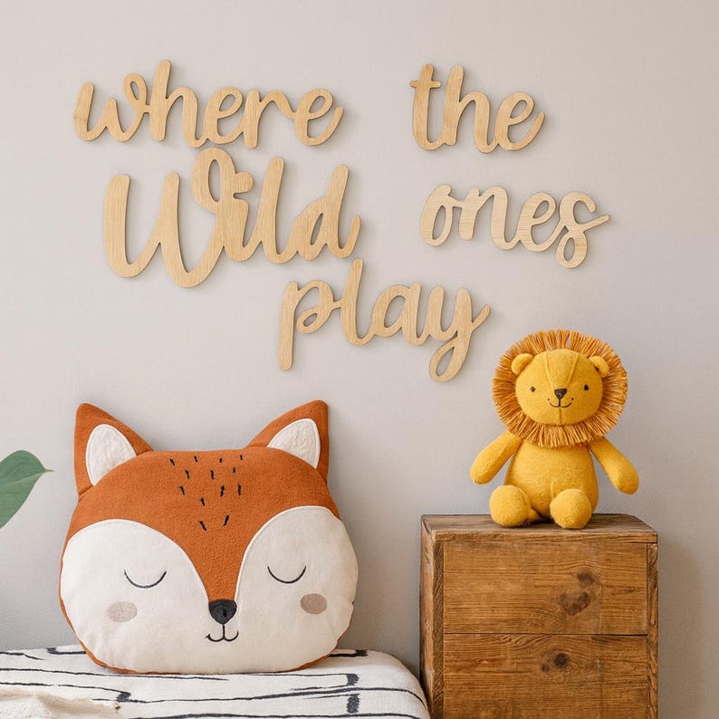 May include: A nursery decor scene featuring a fox-shaped pillow, a plush lion toy, and wooden wall art that reads "where the wild ones play." The fox pillow is orange, white, and beige. The lion is yellow. The wooden letters are light brown.