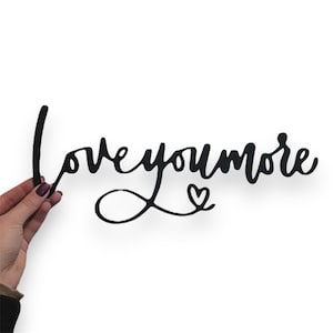 Puede incluir: Arte de pared de metal negro con la frase "love you more" en fuente cursiva. El diseño incluye un bucle que conecta "love" y "you", con un corazón debajo de "you".