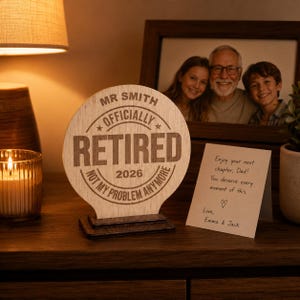 Könnte beinhalten: Eine Holz-Ruhestands-Plakette mit den Worten "MR SMITH OFFICIALLY RETIRED 2026 NOT MY PROBLEM ANYMORE" wird auf einem Holzständer angezeigt. Ein gerahmtes Foto und eine handschriftliche Notiz befinden sich im Hintergrund.