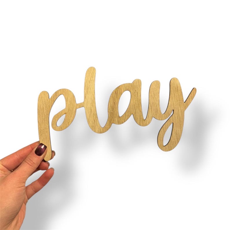 May include: Wooden sign in a cursive font spelling the word "play". The sign is made of light-colored wood and is held by a hand with a dark maroon nail polish. The background is white.