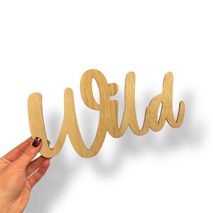 May include: Wooden sign in a cursive font spelling the word "Wild". The sign is made of light-colored wood and is held by a hand with a dark nail polish. The background is white.