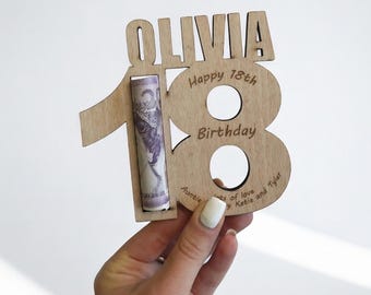 May include: A wooden birthday gift with the name "Olivia" at the top and the number "18" in the center. The number holds a rolled-up bill. The text "Happy 18th Birthday" and "Auntie's of love by Katie and Tyler" are engraved on the wood.