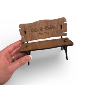 Könnte beinhalten: Eine kleine Holzbank mit dunkelbraunem Finish. Die Bank hat eine geschwungene Rückenlehne und eine Sitzfläche, mit den Namen "Kate & Robin" und dem Datum "22/01/2025" auf der Rückseite eingraviert. Die Bank wird von einer Hand gehalten.