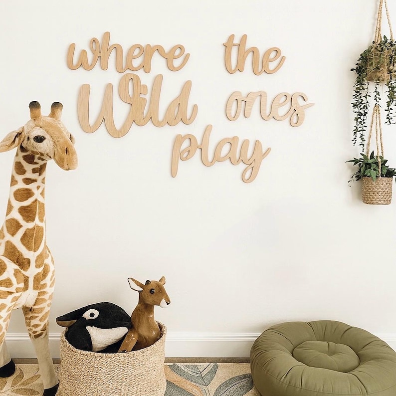 May include: A nursery scene with a giraffe plush toy, a woven basket with stuffed animals, and wooden wall art that reads "where the wild ones play." Hanging plants and a green beanbag chair complete the room's decor.