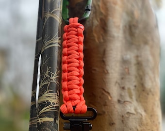 May include: A bright orange paracord sling with a black swivel clip attached to a camouflage shotgun.