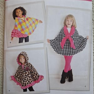 May include: Simplicity sewing pattern 1706A featuring children's capes. The pattern includes capes with a plaid design, a black and white houndstooth pattern, and a leopard print. The capes have hoods and ruffled edges. The pattern size is S-L.