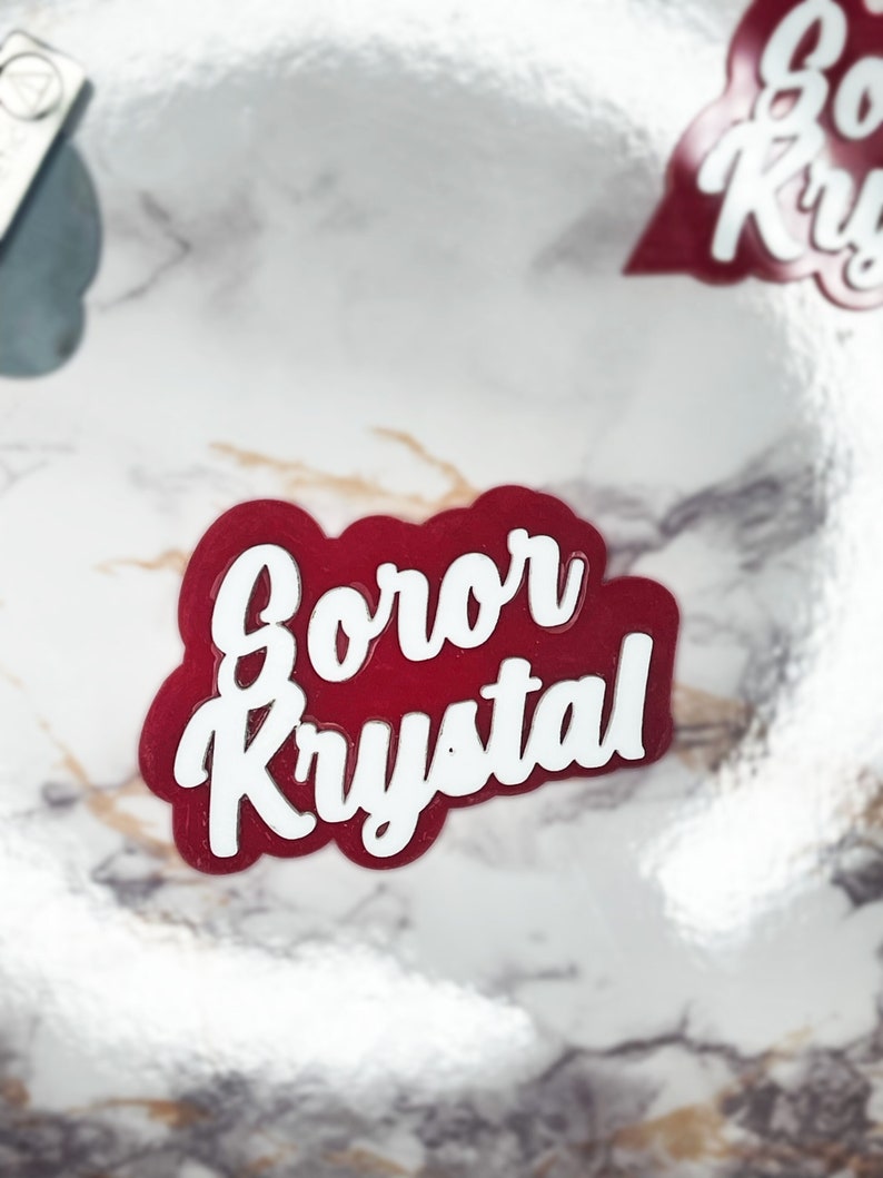 May include: A red and white sticker with the text "Gorer Krystal" in a cursive font. The sticker has a rounded shape and is set against a marble background. Another sticker is visible in the background.
