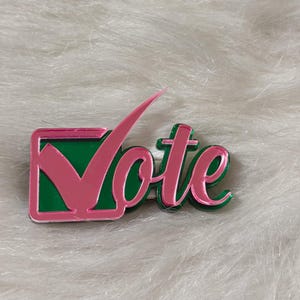 May include: A pink and green acrylic pin with the word "Vote" and a checkmark inside a square.