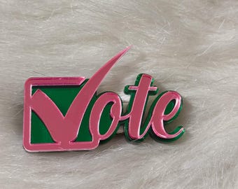 May include: A pink and green acrylic pin with the word "Vote" and a checkmark inside a square.