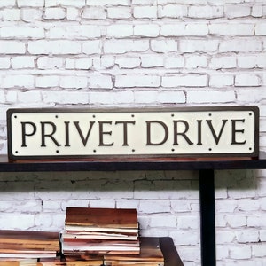 May include: A white sign with black lettering that reads "PRIVET DRIVE". The sign is made of wood and has a distressed look. It is sitting on a shelf against a white brick wall.