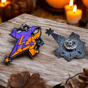Pode incluir: Dois pinos de esmalte, um com um design geométrico roxo e laranja e o outro com um design de glitter prateado. Ambos os pinos têm uma estrela no topo e uma parte de trás do pino.