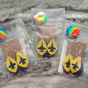 May include: Three clear plastic bags with yellow and blue leather earrings shaped like airplanes. Each bag has a small round sticker with the words "Thank You" printed on it.