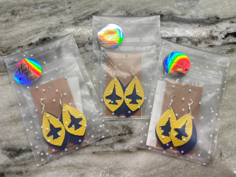 May include: Three clear plastic bags with yellow and blue leather earrings shaped like airplanes. Each bag has a small round sticker with the words "Thank You" printed on it.