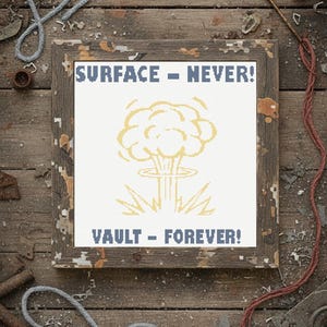 May include: Framed cross-stitch art with a distressed wooden frame. The artwork features a yellow mushroom cloud design with the text "SURFACE - NEVER!" and "VAULT - FOREVER!" in blue. The background is white.