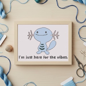 May include: A framed cross-stitch featuring a light blue cartoon creature with the text "I'm just here for the vibes." The artwork is surrounded by blue thread, spools, a thimble, and scissors on a light wood surface.