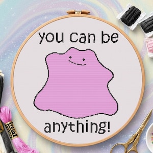 May include: A cross-stitch hoop featuring a pink blob character with a smiling face. The text "you can be anything!" is stitched above and below the character in black thread. The hoop is set in a wooden frame, with embroidery floss and scissors in the background.
