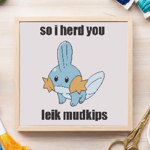 May include: A framed cross-stitch featuring a blue cartoon creature with orange accents. The text "so i herd you" and "leik mudkips" are stitched above and below the character. The frame is light-colored wood.