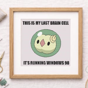 May include: Framed cross-stitch art with a light wood frame. The artwork features a cartoon brain cell with the text "THIS IS MY LAST BRAIN CELL IT'S RUNNING WINDOWS 98". Embroidery floss, scissors, and a pincushion are also visible.