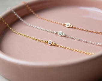 May include: Three delicate necklaces with a single small clear stone charm on each. The necklaces are on a pink surface and are in gold, silver, and rose gold.
