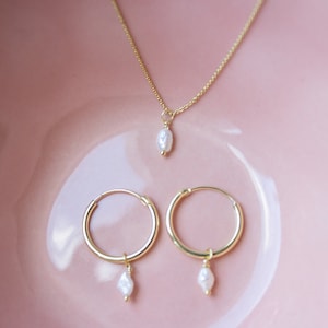 Könnte beinhalten: Ein Set aus goldenen Creolen und einer goldenen Halskette mit einem einzelnen Perlenanhänger. Die Ohrringe und die Halskette liegen auf einer rosa Oberfläche.